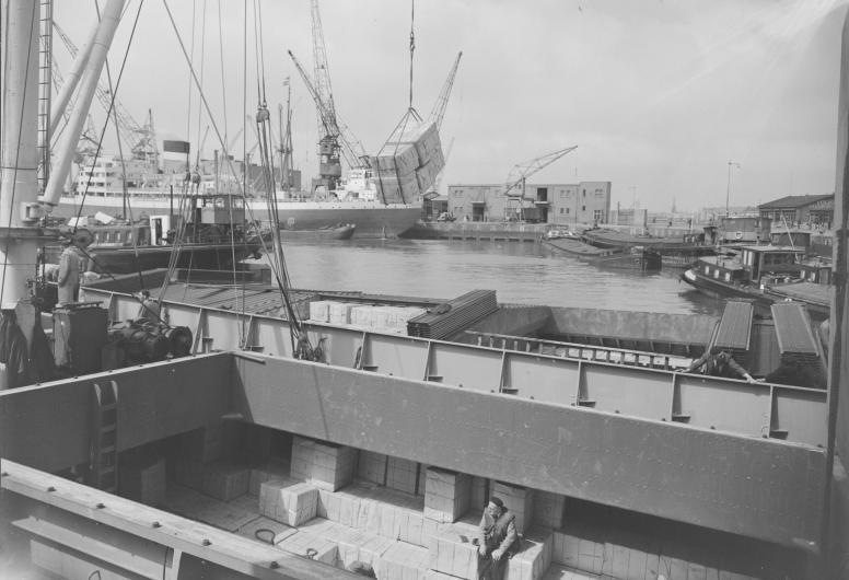 Overslag van stukgoederen in de Merwehaven. Het laden en lossen van goederen vanuit een het ruim van een schip naar een binnenvaartschip (1953).