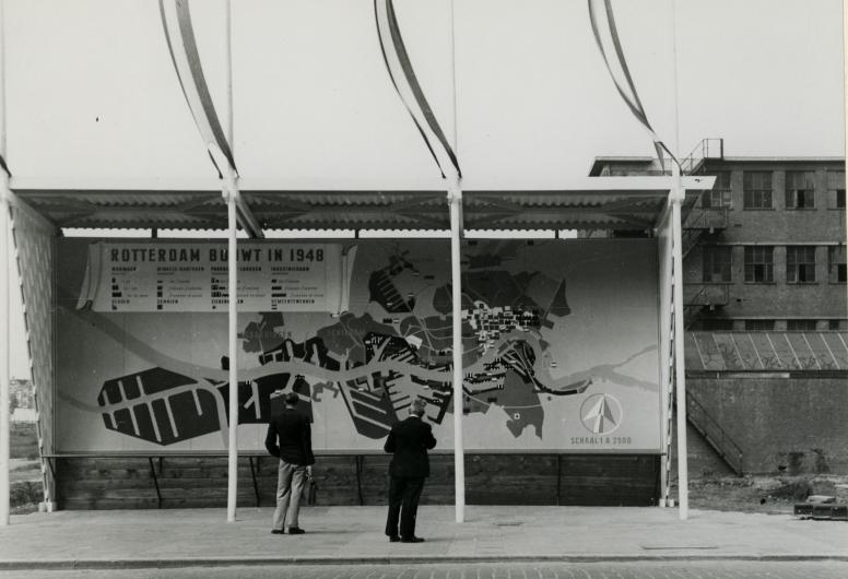 Twee mensen bekijken een groot promotiebord 'Rotterdam Bouwt in 1948' op de Coolsingel. Op de achtergrond rechts een deel van de Ravero garage, 1948. Collectie 4135, XXVI-49-00-00-01