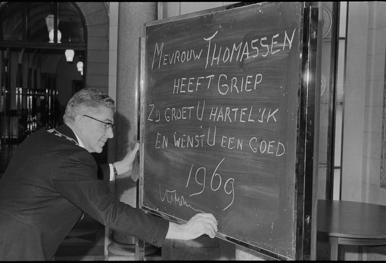 1 januari 1969: Burgemeester Thomassen schrijft met krijt op een bord dat mevrouw Thomassen griep heeft. Ondanks dat zij niet aanwezig is bij de traditionele nieuwjaarsreceptie in de burgerzaal van het stadhuis, wenst de burgemeestersvrouw iedere bezoeker van de receptie een 'goed 1969'. Fotograaf Ary Groeneveld. Collectie 4121, 21225-1-2-kl