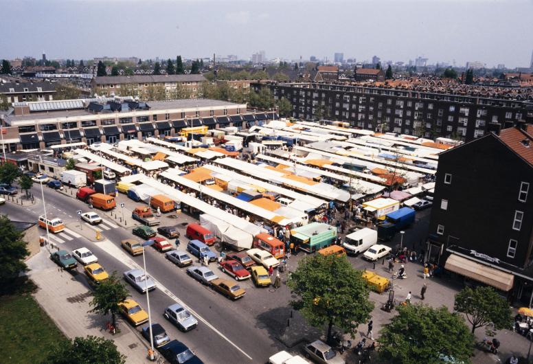 Zicht op de zaterdagmarkt op het Visserijplein, gezien vanaf de bovenste verdieping van de flat op de hoek van de Gijsinglaan en de Grote Visserijstraat. Links ligt gebouw van het Openbare Bibliotheekfiliaal Delfshaven. 23 mei 1987.
