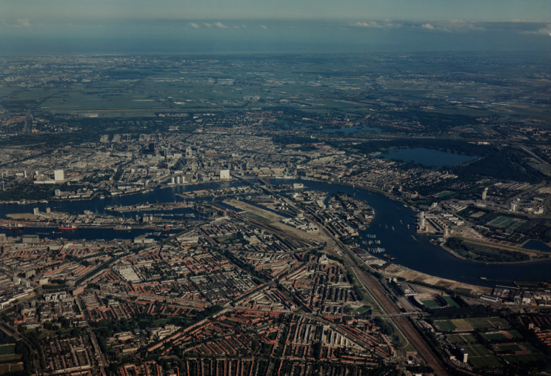1991-3348: Luchtopname van Rotterdam vanuit het zuiden, met op de voorgrond Feijenoord, in het midden de Nieuwe Maas en op de achtergrond de Stadsdriehoek. Rechts de wijk Kralingen en daarachter de Kralingse Plas en het Kralingse Bos, en op de achtergrond