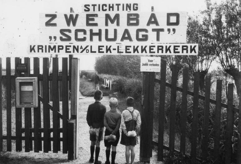 Drie jongens bij het hek van Zwembad Schuagt te Krimpen a/d Lek - Lekkerkerk met daarop een bord met de tekst: Voor Joden verboden. Foto Johannes van Rhijn. Collectie 4140, nr. 1977-3206